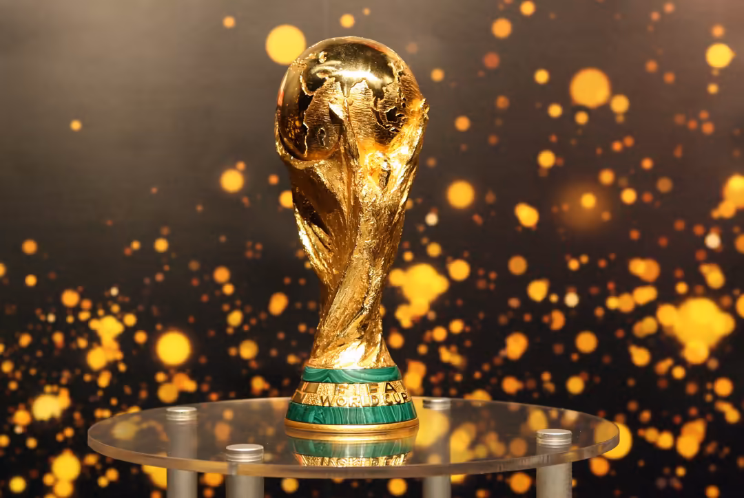 Golden FIFA World Cup trophy on display pedestal with shimmering golden bokeh particles and dark background creating celebrat