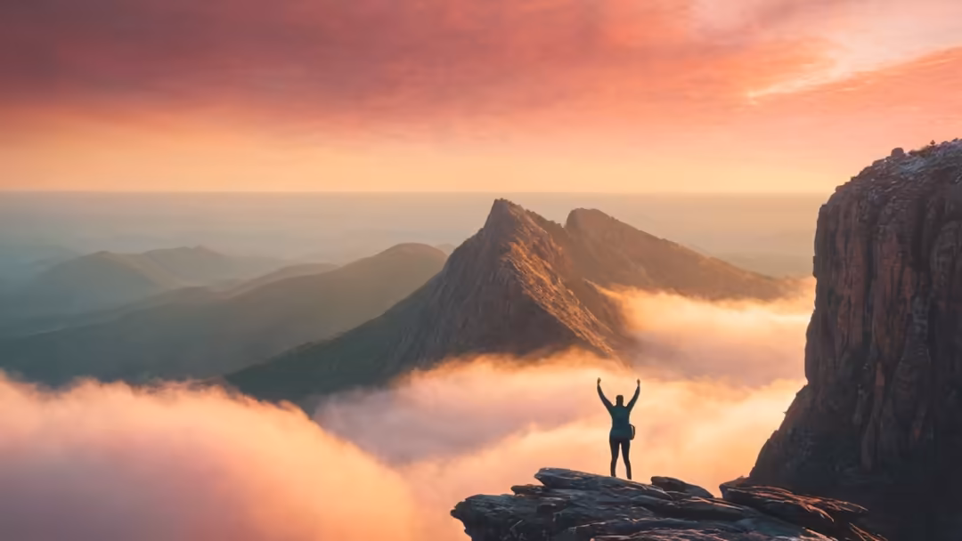 Person standing on rocky cliff with arms raised in triumph, overlooking misty mountains at golden sunrise with warm peachy sk