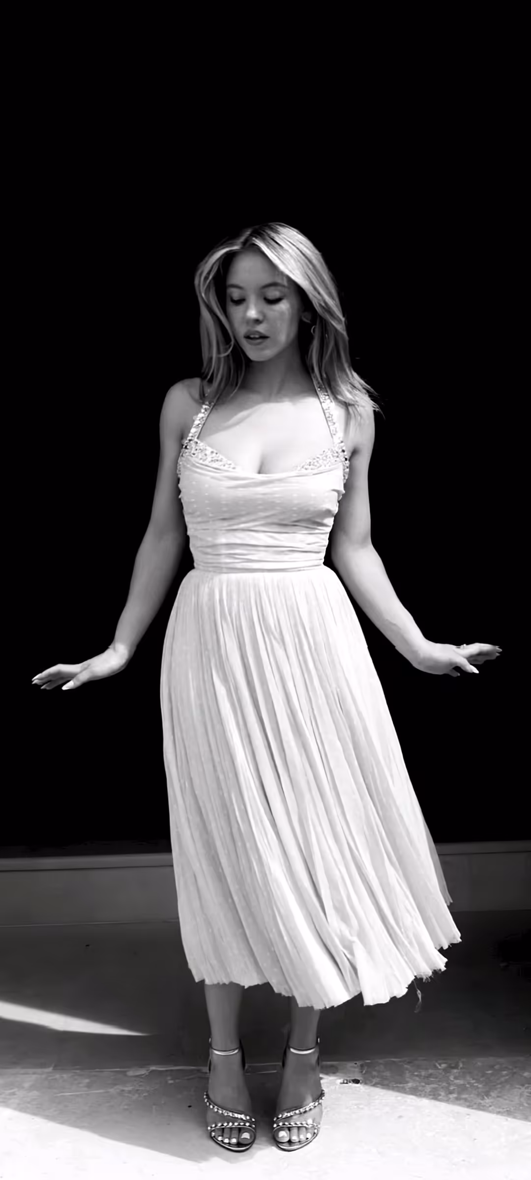 Sydney Sweeney in elegant white pleated dress against dramatic black background, black and white portrait photography with so