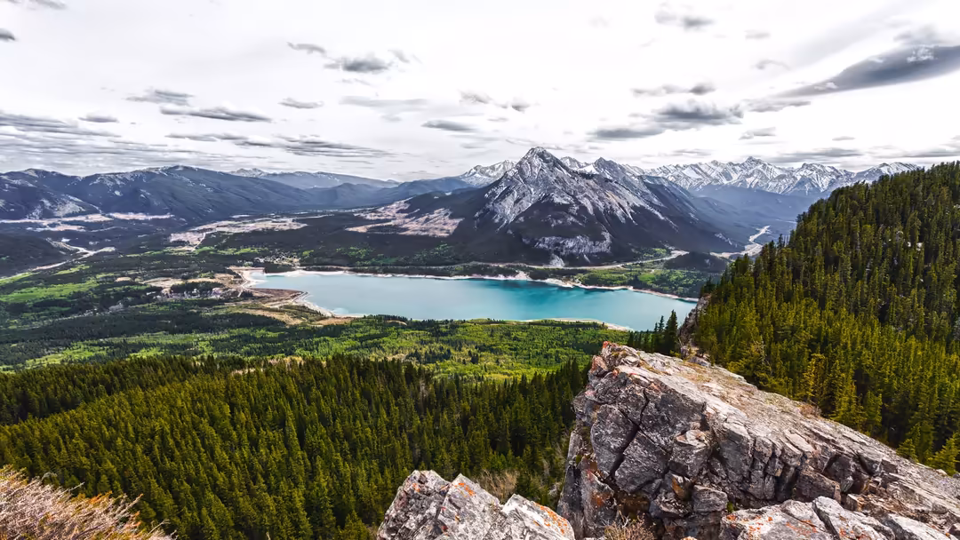 Turquoise alpine lake nestled in a green mountain valley with snow-capped peaks and dense pine forests under dramatic cloudy