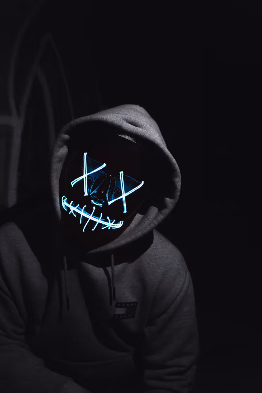 Hooded figure in darkness wearing glowing blue neon mask with X markings and stitched mouth design, cyberpunk aesthetic