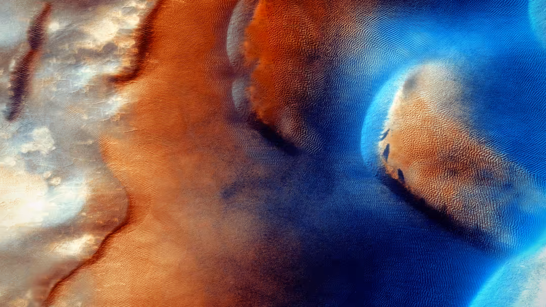Close-up of Mars surface with rust-orange and cream-colored terrain features, blue atmospheric glow creating an otherworldly,