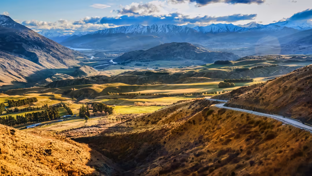 Sweeping mountain valley landscape with snow-capped peaks in distance, winding river, golden fields, and brown hillsides unde