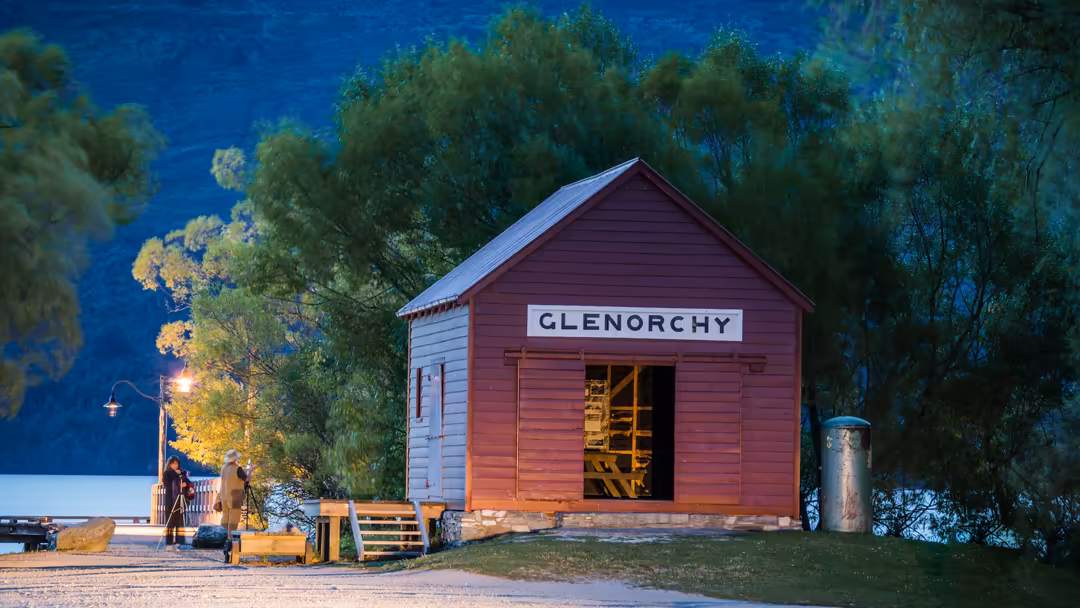 Red and white wooden station building labeled Glenorchy situated by a deep blue lake, surrounded by lush green trees at dusk
