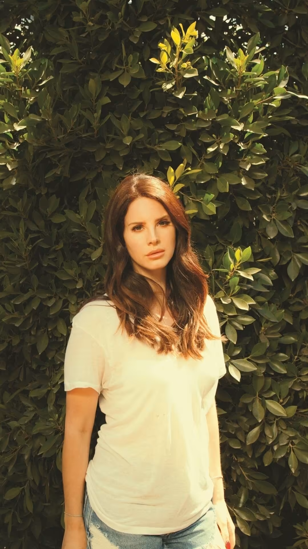 Woman with auburn hair wearing white shirt posing against lush green foliage with yellow flowers in natural sunlight