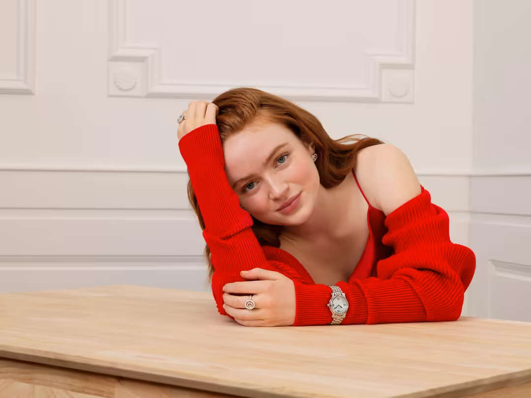 Woman with reddish-brown hair wearing a bright red off-shoulder sweater, leaning on a wooden table against a white decorative