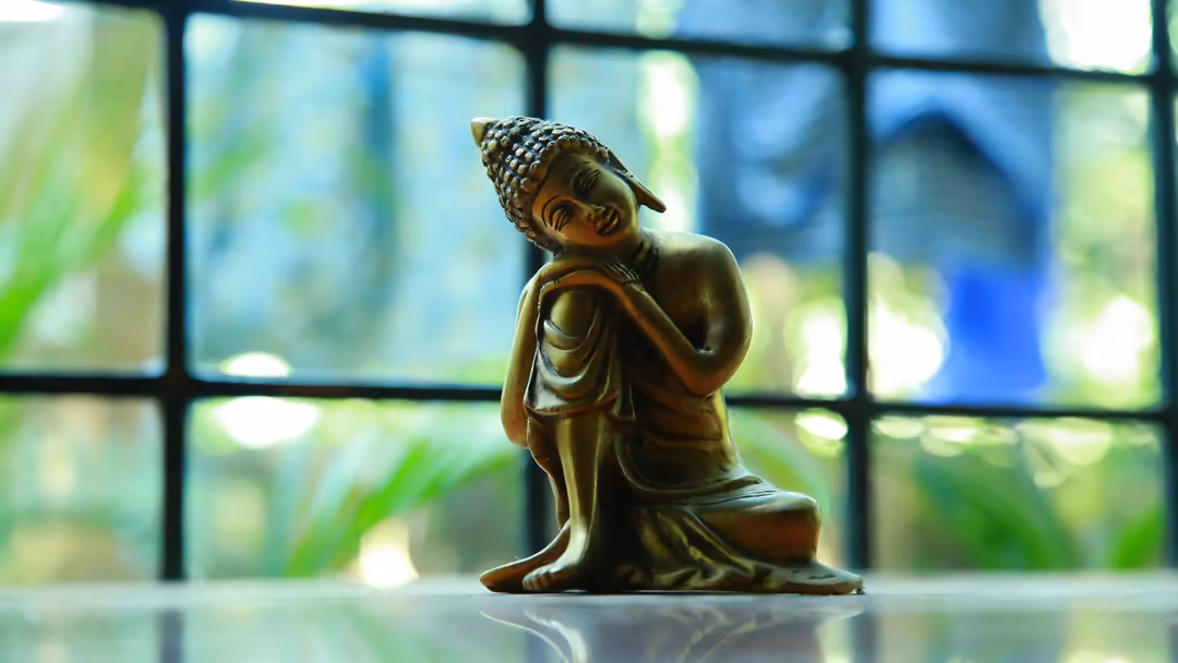 Bronze Buddha statue in serene meditation pose displayed on reflective surface with blurred garden window background