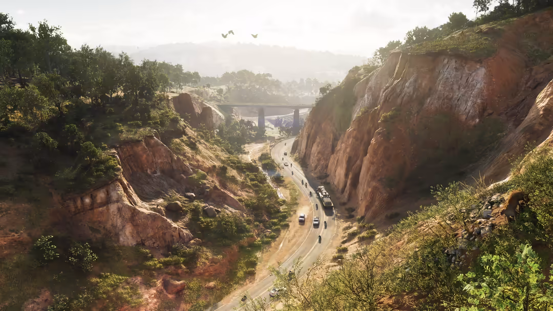 Aerial view of a winding road through a deep canyon with red rocky cliffs, lush green vegetation, a distant bridge, and vehic