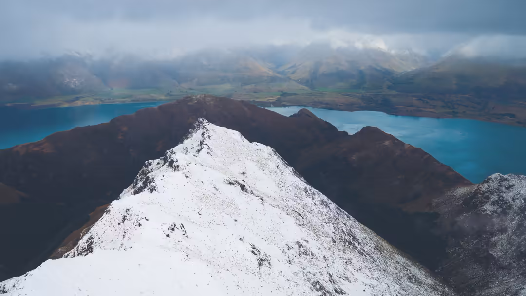 Snow-covered mountain peak overlooking vibrant turquoise alpine lakes surrounded by dramatic peaks and cloud-covered valleys