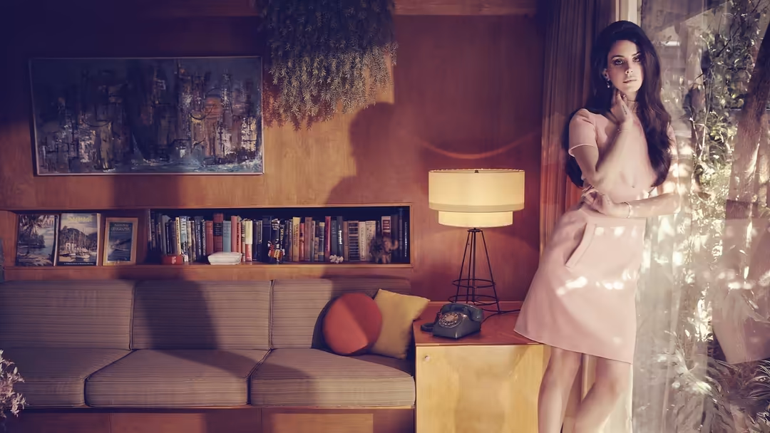 Woman in pale pink dress standing in warm-toned vintage living room with bookshelves, lamp, and hanging plants by window