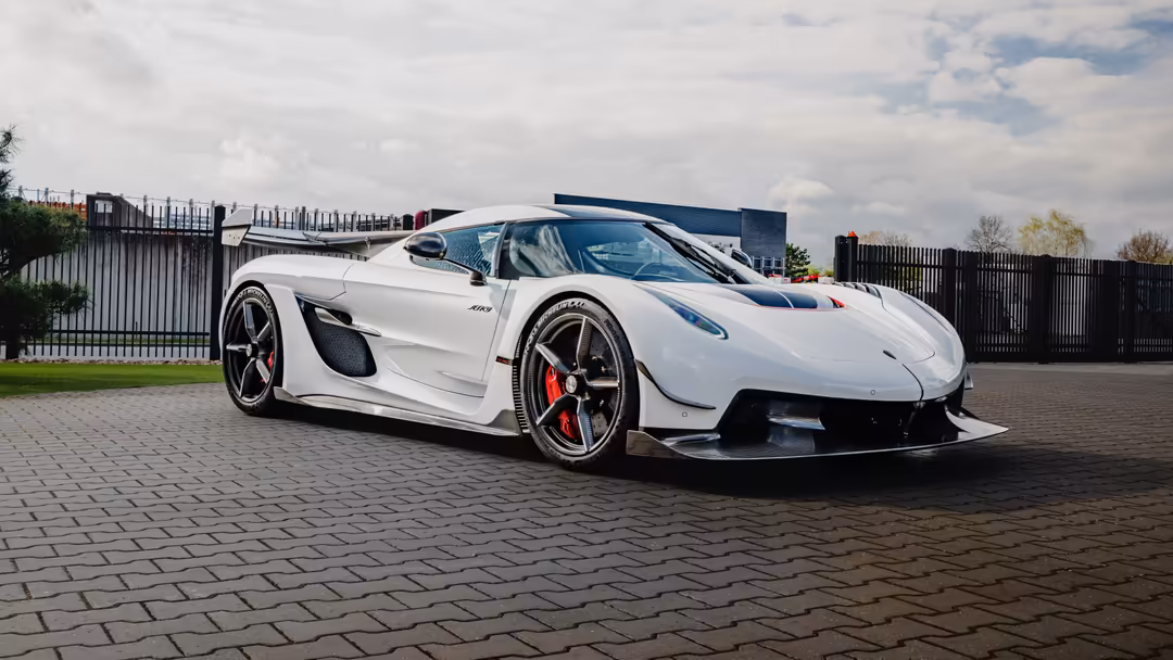 White Koenigsegg Jesko hypercar parked on paved surface with industrial fencing and cloudy sky in background