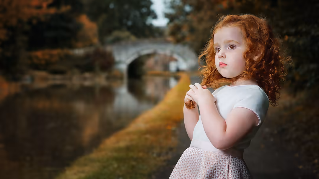 Young child with curly red hair wearing white shirt and pink skirt, posing thoughtfully in front of blurred vintage rural set