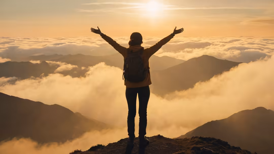 Silhouette of hiker with arms raised triumphantly atop mountain peak overlooking sea of golden clouds at sunrise
