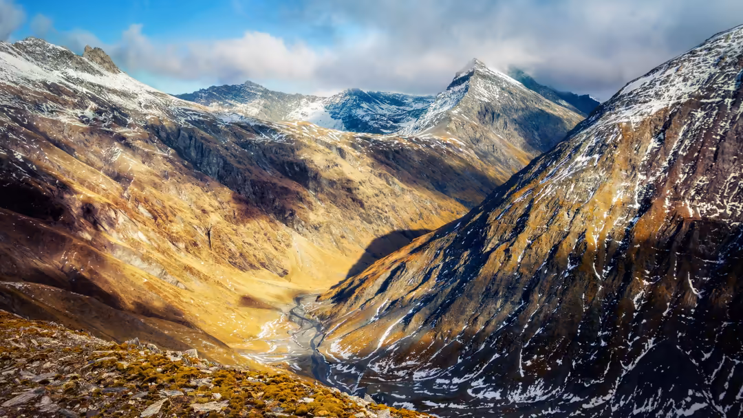 Majestic snow-capped mountain range with golden-brown valleys and a winding river cutting through the alpine landscape under