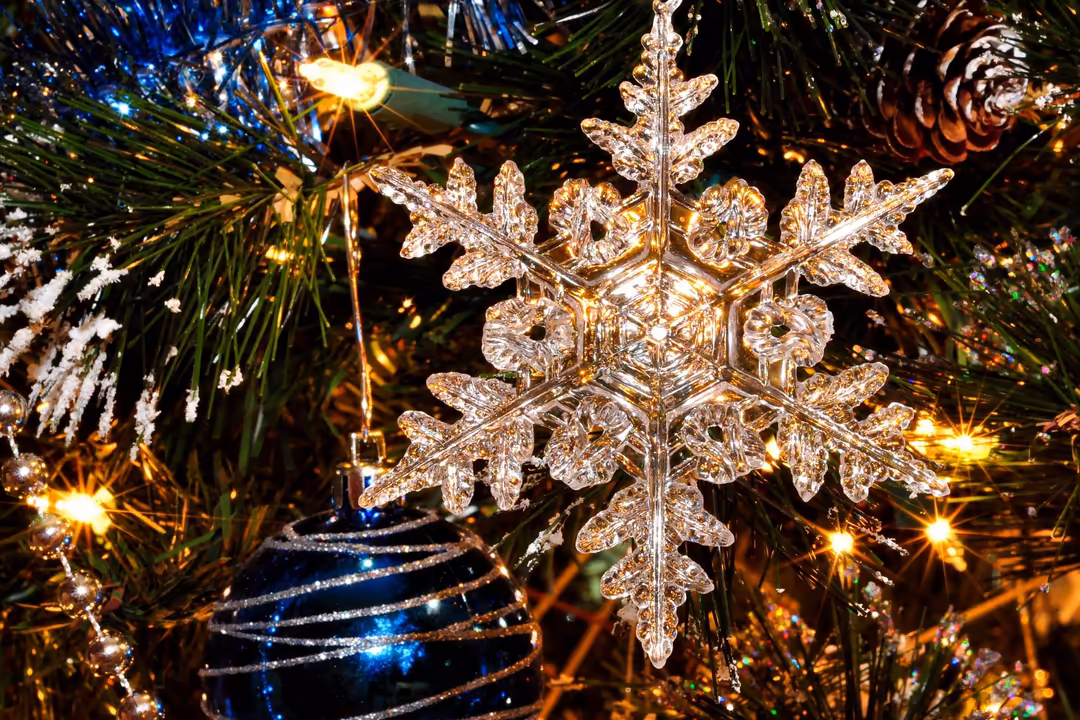 Crystal snowflake ornament with golden sparkle against blue holiday lights and evergreen branches with pinecones