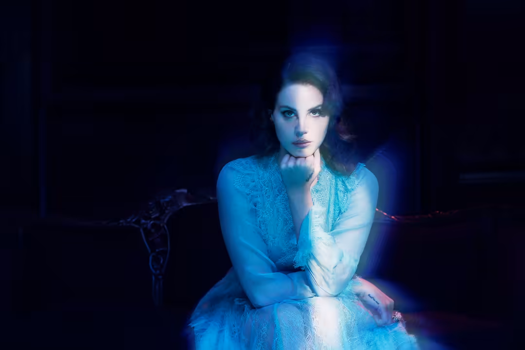 Woman with dark hair in blue lace dress seated against dark background with ethereal blue glow lighting her face and shoulder