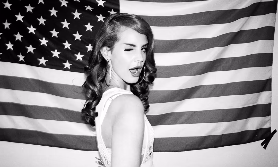 Black and white portrait of Lana Del Rey against American flag backdrop, wearing white top with vintage wavy hair styling and