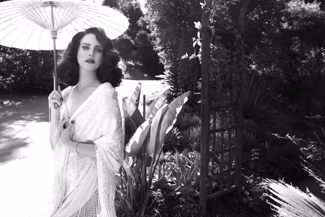 Black and white portrait of woman in white robe holding parasol in lush garden with ivy-covered arbor and tropical plants