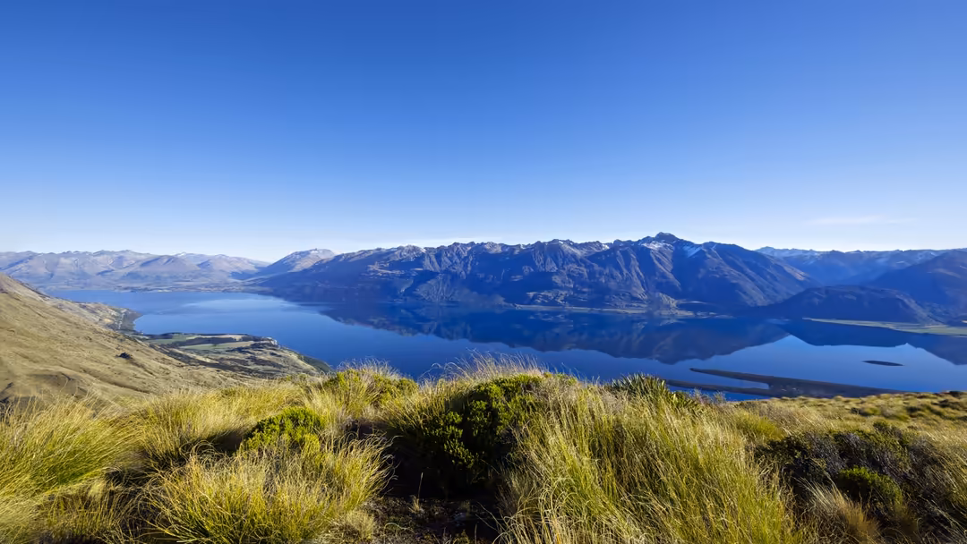 Expansive alpine valley with deep blue lake surrounded by dramatic mountain peaks under clear sky, lush green grassland in fo
