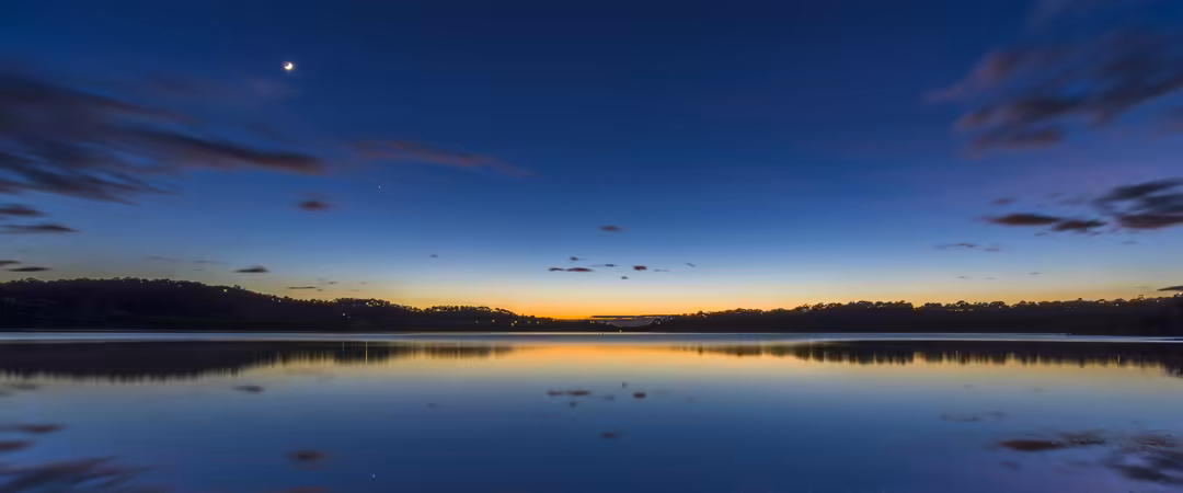 A serene lake at twilight with perfect mirror reflections of the golden sunset sky, moon rising, and forested shoreline under