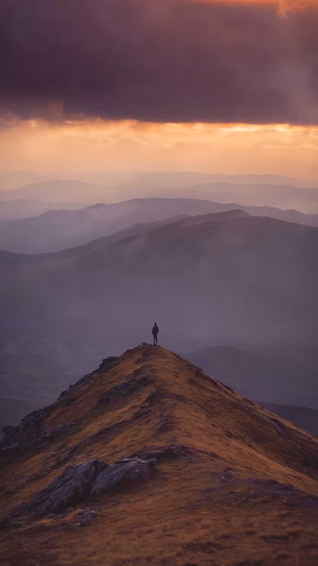 Solitary figure standing on rocky mountain peak at sunset, dramatic layered mountain ranges receding into misty distance unde