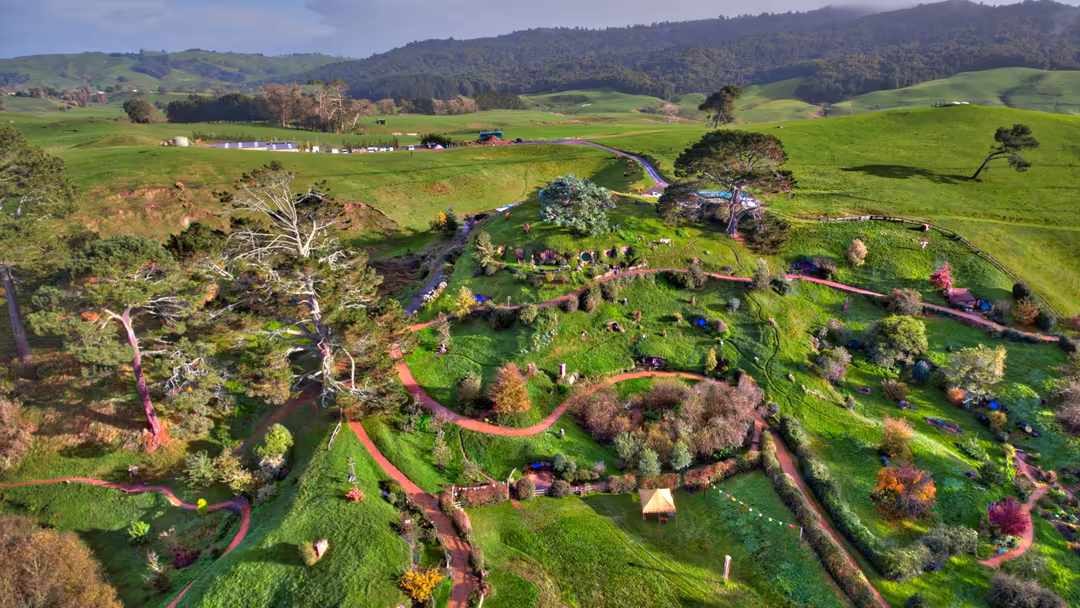 Aerial view of lush green rolling hills with a winding river, scattered farmhouses, trees, and vibrant flowering gardens dott