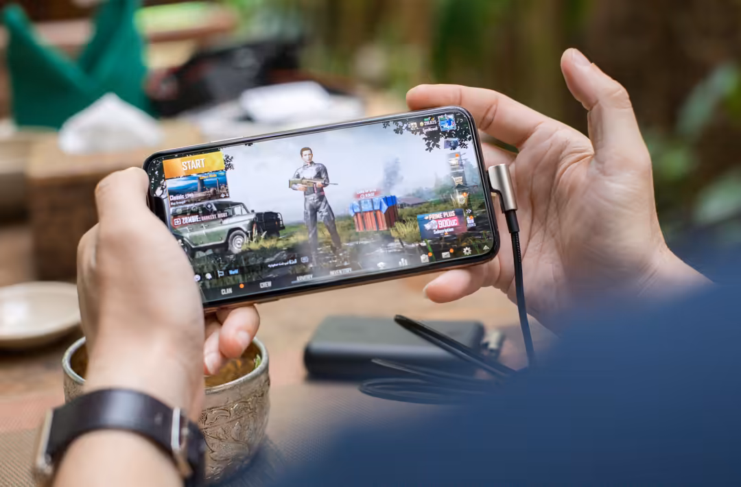 Hands holding smartphone displaying PUBG mobile game with player and vehicles on screen, outdoor environment blurred in backg