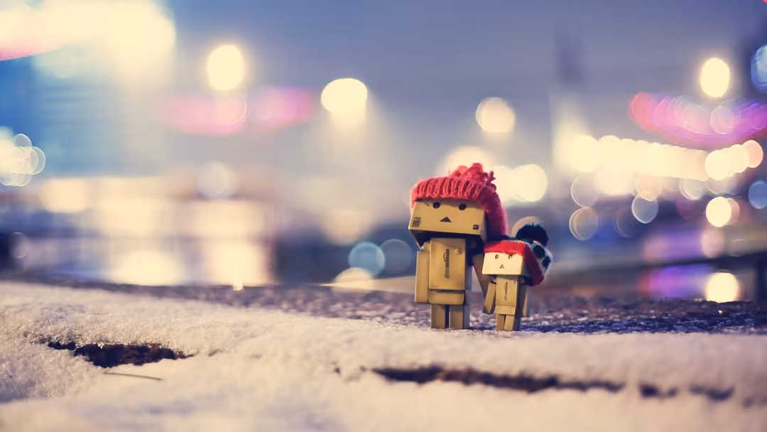 Two cardboard box figures wearing red knit hats stand on snowy ground with soft bokeh city lights glowing in the blurred back