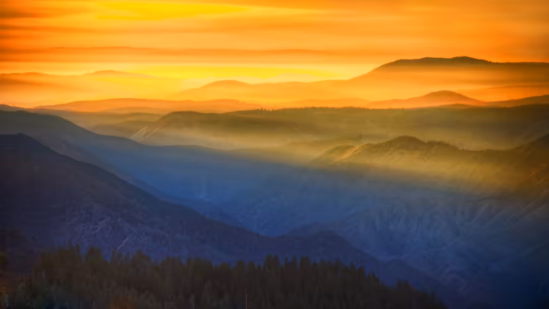Golden sunset illuminates layered mountains and misty valleys with warm orange and blue tones, forested peaks in foreground s