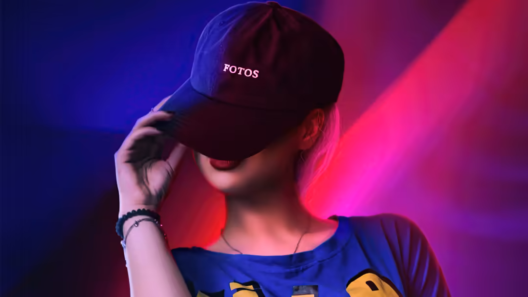 Person wearing black cap and blue shirt posed against vibrant neon pink and blue gradient backdrop with atmospheric lighting