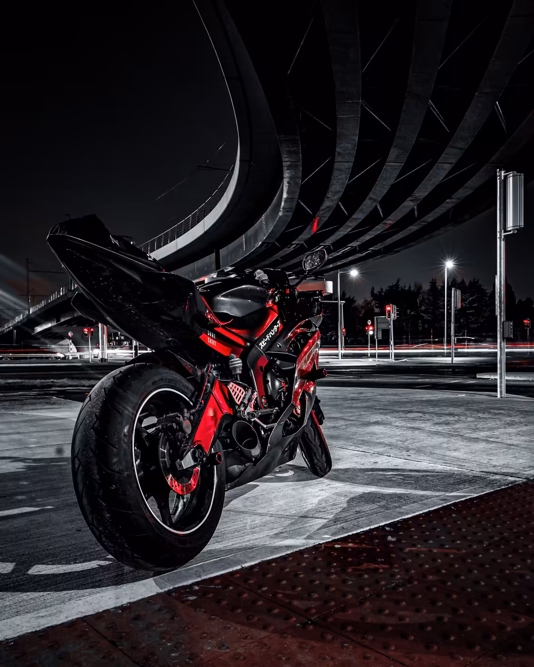Red motorcycle parked under curved highway overpass at night with city lights and dark sky in background