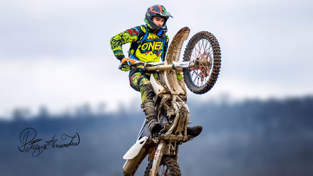 Motocross rider in neon yellow gear performing aerial wheelie on dirt bike against blurred forest background