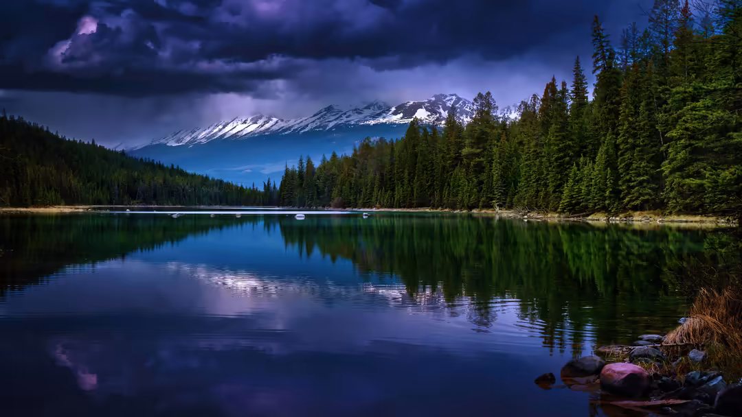 Snow-capped mountain reflected in calm alpine lake surrounded by dense evergreen forest with dramatic purple storm clouds ove