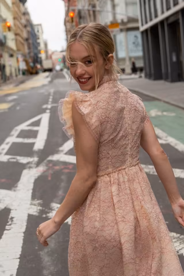 Sydney Sweeney in a pink lace dress smiling over her shoulder on a sunny urban street with buildings and yellow traffic light