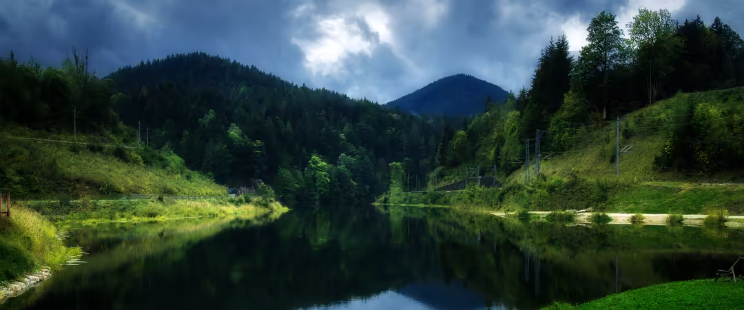 Serene mountain valley with lush green forests, a calm reflective river, rolling hills, and dramatic sky with clouds overhead