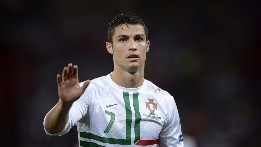 footballer in white Portugal national team jersey with green and red stripes raises hand against dark background.