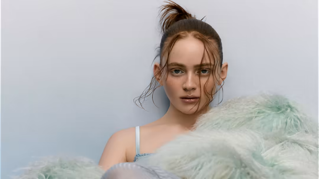 Young woman with reddish-brown hair in bun, wearing light blue strap and green tulle, gazing directly at camera against neutr