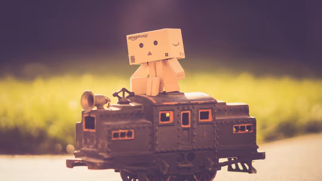 Adorable cardboard box robot with smiling face sitting atop a vintage toy train against a soft, blurred yellow and green back