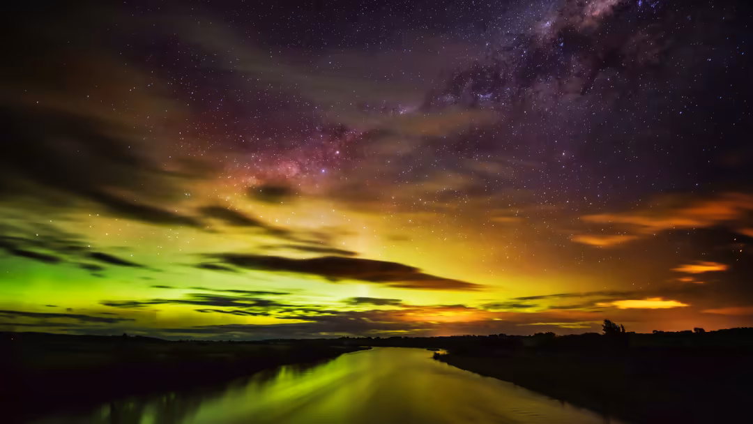Vibrant aurora borealis dancing across starlit sky with green and purple hues reflected in calm river below at night