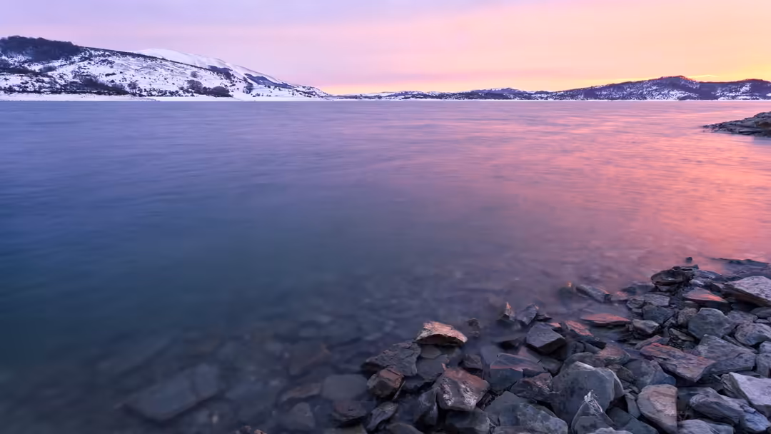 Snow-covered mountains reflected in a calm frozen lake during a dramatic purple and pink sunset with rocky foreground