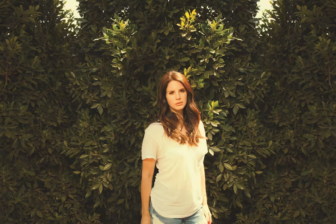 Woman with reddish-brown hair in white shirt standing against lush green foliage backdrop with natural lighting
