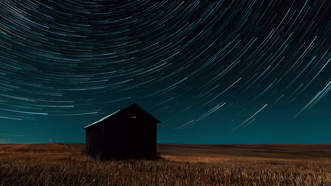 Abandoned rural house silhouetted against a teal night sky with long exposure star trails and streaking constellations overhe