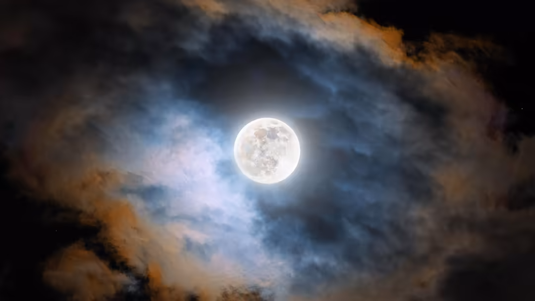 Bright full moon glowing through dark stormy clouds with blue and orange atmospheric lighting creating a dramatic nighttime s