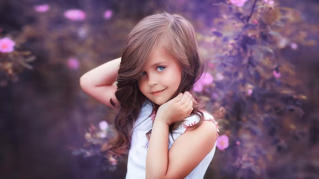 Young girl with long brown hair posing gracefully among purple flowers in a dreamy, soft-focused aesthetic setting
