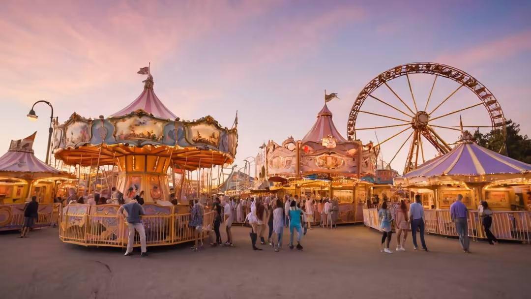 Vibrant theme park at sunset with illuminated carousel, ferris wheel, and pink-striped tents filled with visitors enjoying th