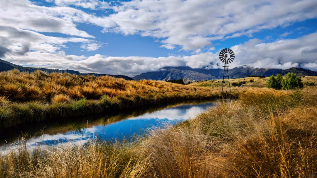 A windmill stands in golden grassland with a blue stream, mountains in distance under dramatic cloudy sky