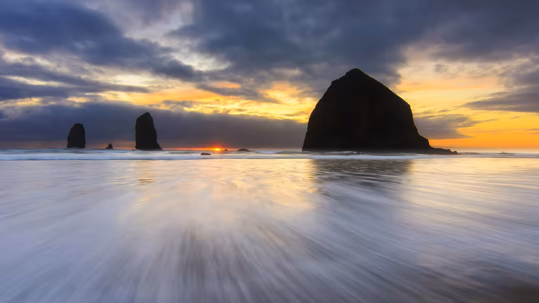 Dramatic coastal landscape with towering dark rock formations silhouetted against a golden sunset sky with orange and blue cl