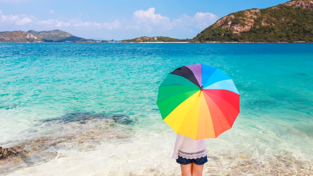Person holding a vibrant rainbow umbrella standing in crystal-clear turquoise water with lush green hills and white sand beac