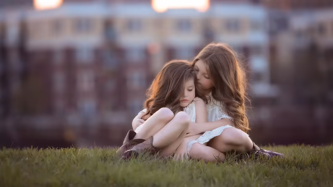Two young girls sitting together on grass in golden sunlight, one kissing the other's cheek in a tender moment of sisterly af