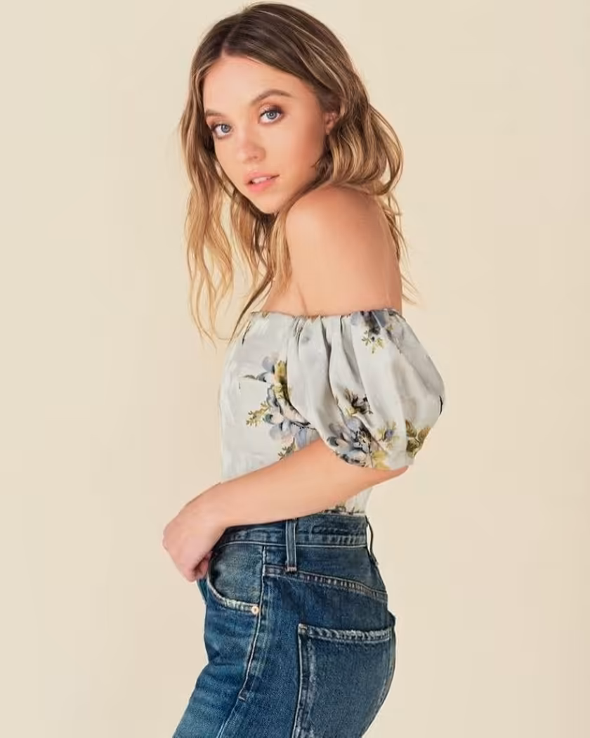 Sydney Sweeney with long wavy brown hair wearing a light cream floral off-shoulder top and blue jeans, posed against a neutra
